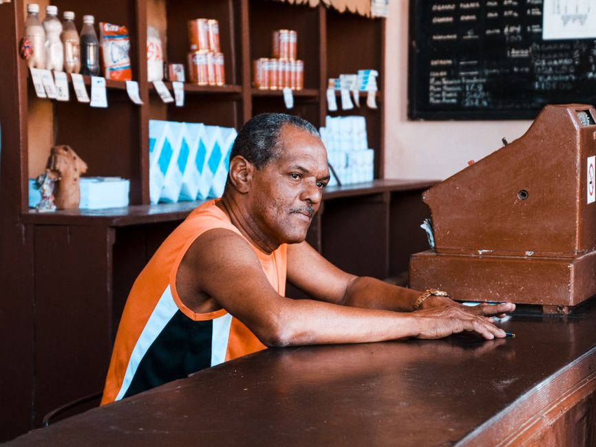 A man in a vintage setting, leaning on a wooden counter beside a cash register in a store.